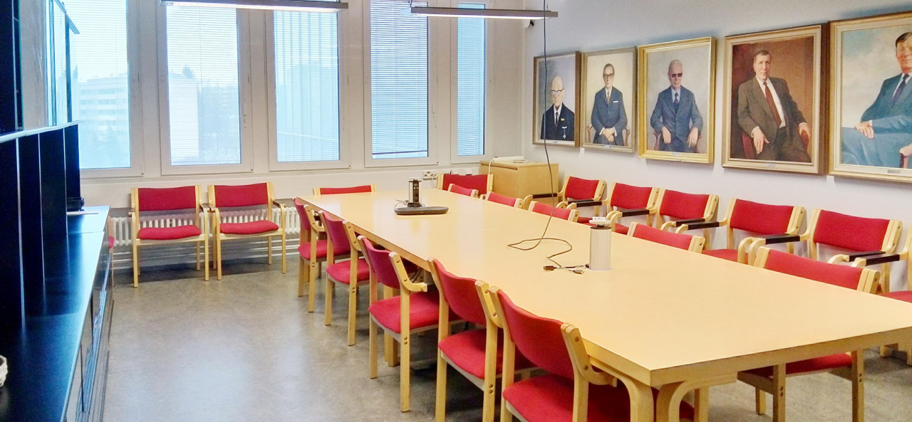 Pitkä pöytä, jonka ympärillä ja seinänvierustoilla punaisia tuoleja. Pöydän keskellä kokoustekniikkaa: johtoja, pistorasioita, kaiutin. Yhdellä seinustalla isot ikkunat, toisella seinällä tauluja, joissa miehiä harmaat puvut yllään.